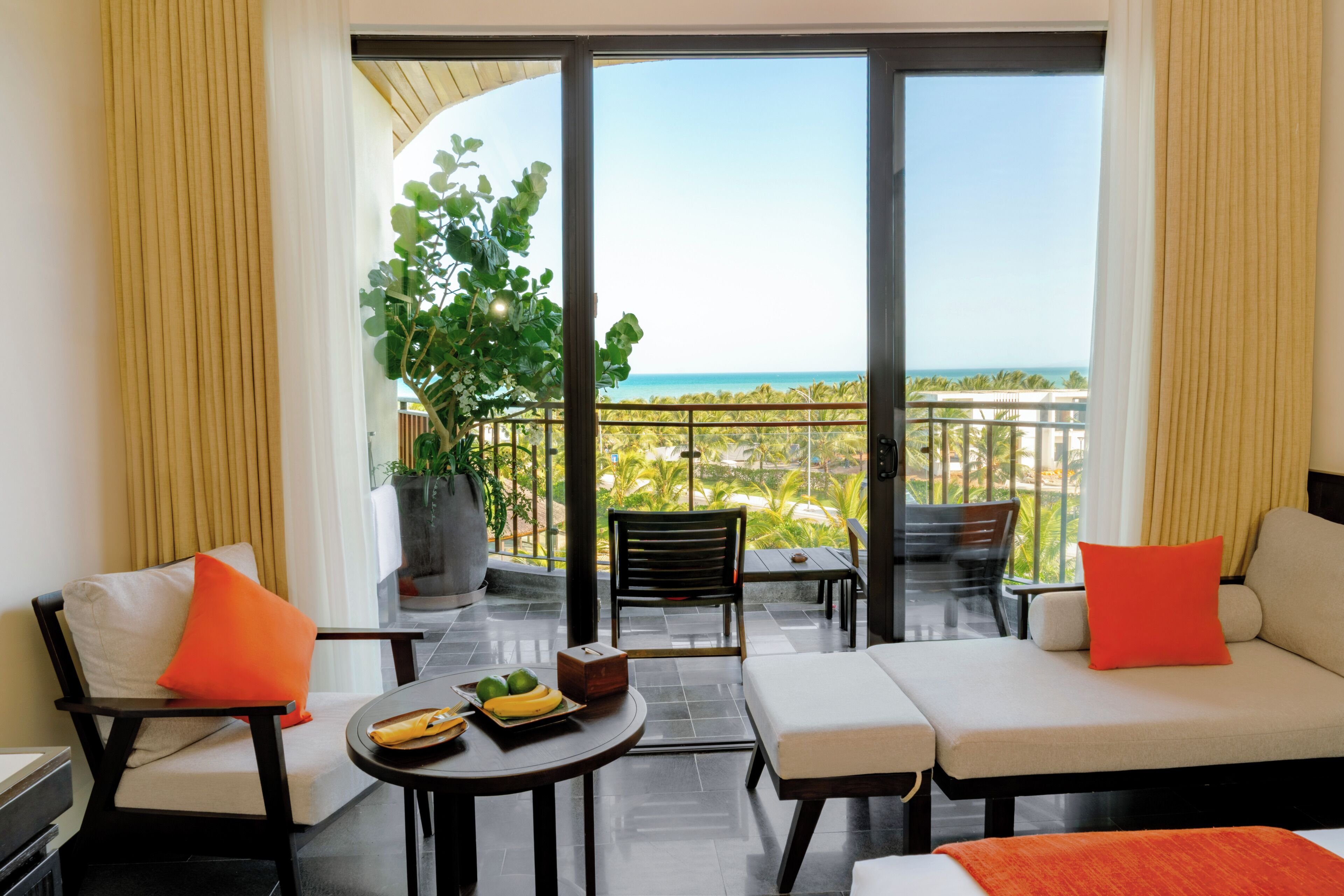 A balcony with a view of the ocean, a sofa, two chairs, a small table with fruit, and a potted plant.