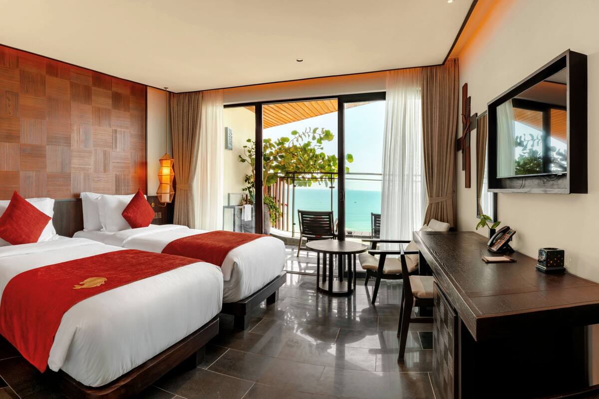 A hotel room with two beds, a desk, a chair, and a balcony with a view of the sea.