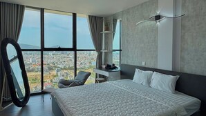 A modern bedroom with a large bed, a chair, a vanity, and a view of a cityscape through large windows.