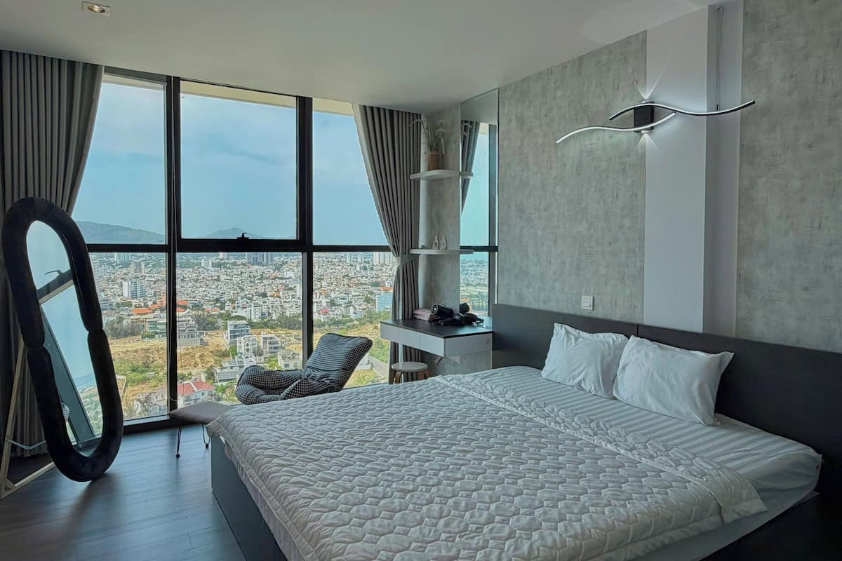 A modern bedroom with a large bed, a chair, a vanity, and a view of a cityscape through large windows.