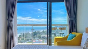 A bedroom with a large window, a bed, a yellow armchair, and a view of a coastal area.