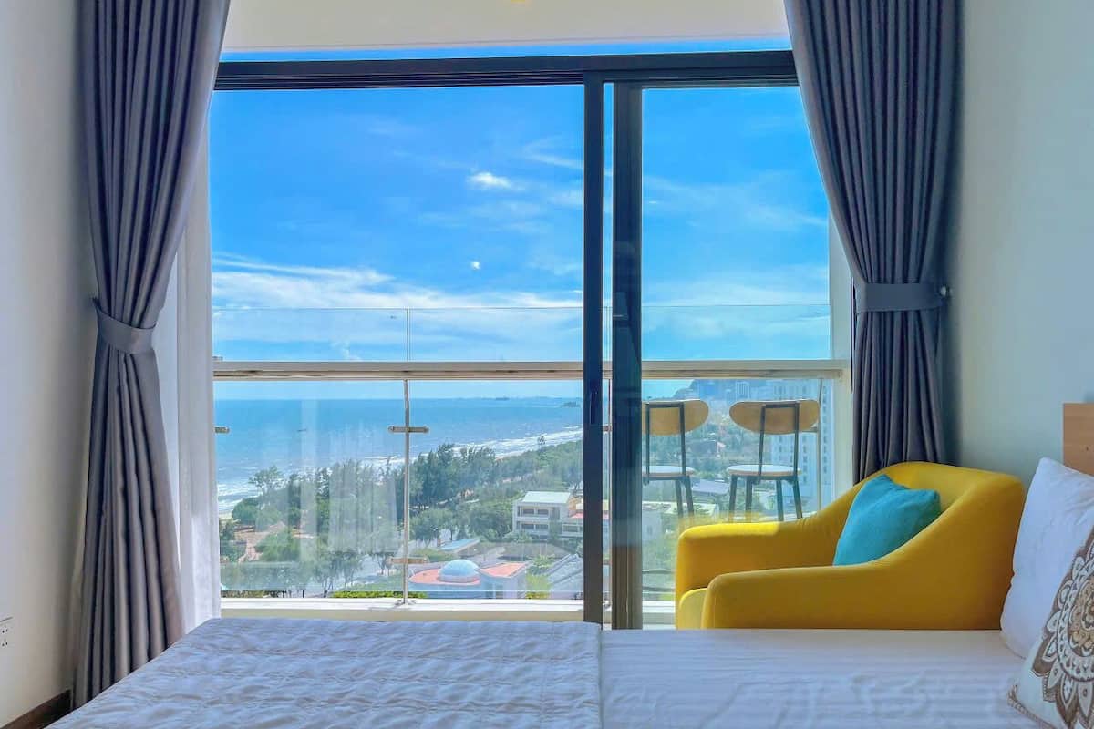 A bedroom with a large window, a bed, a yellow armchair, and a view of a coastal area.