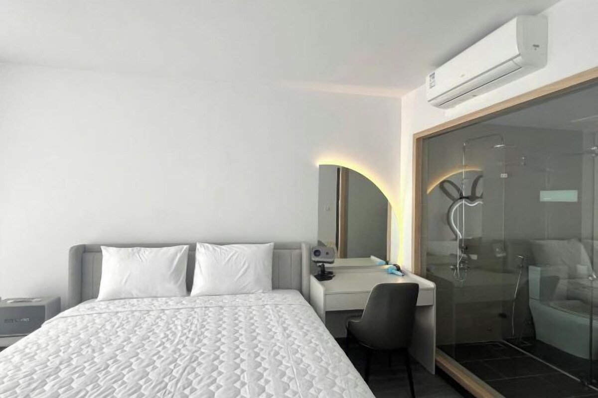 A modern hotel room with a bed, a desk, a chair, a mirror, and a shower area.