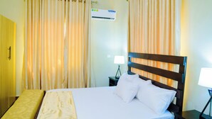 A hotel room with a bed, curtains, an air conditioning unit, and a bedside table with a lamp. - Consulate Hotel (Ibadan)
