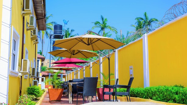 A row of outdoor tables and chairs with umbrellas, yellow walls, and palm trees in the background. - Consulate Hotel (Ibadan)