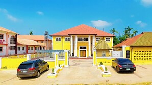 A yellow building with a red roof, two cars parked in front, and palm trees in the background. - Consulate Hotel (Ibadan)