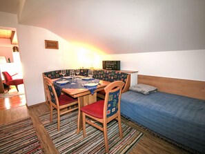 Um quarto pequeno e acolhedor com uma mesa de madeira posta para refeições, uma cama simples com um cobertor azul e um tapete colorido no chão.