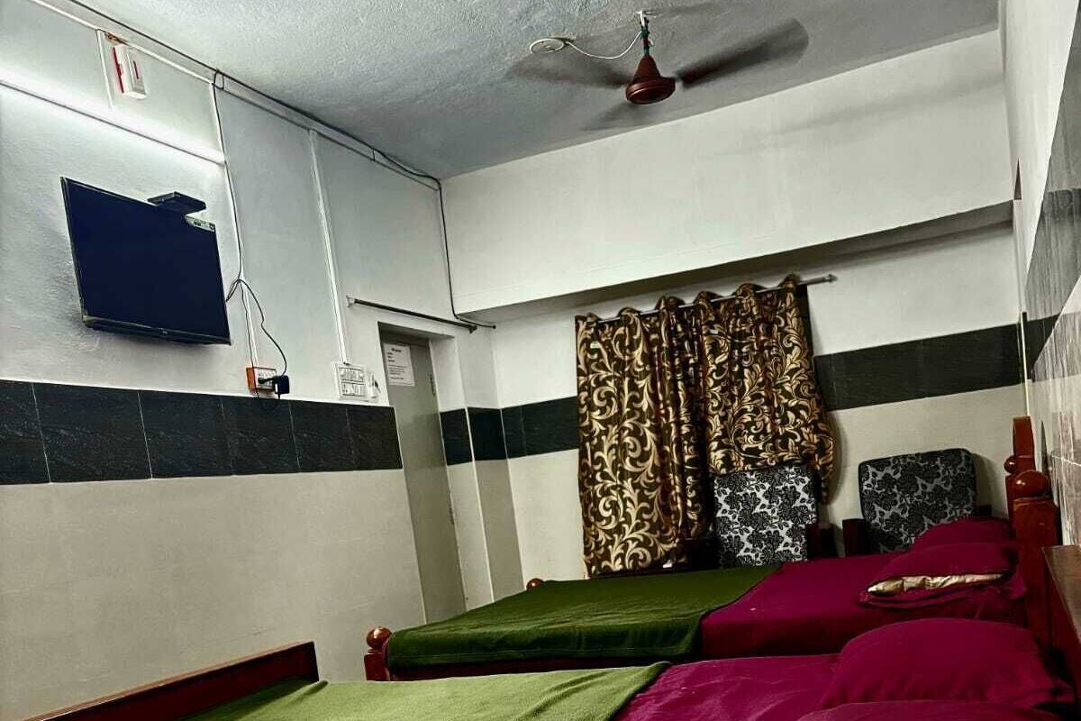 A room with two single beds, a ceiling fan, a television mounted on the wall, and a patterned curtain.