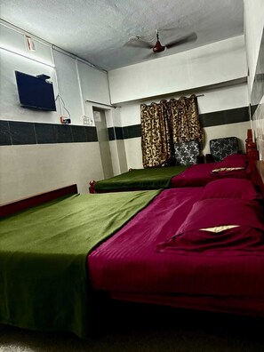 Une chambre avec deux lits simples, un ventilateur de plafond, un téléviseur fixé au mur et un rideau à motifs.
