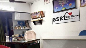 A small office with a desk, a chair, and a computer. There is a wall-mounted shelf with a small statue and a framed picture. A 'Stay Home' sign is visible on the wall. - GSR Stay Home (Puducherry)