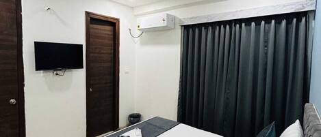 A hotel room with a bed, a ceiling fan, an air conditioning unit, a TV, and a window with curtains.