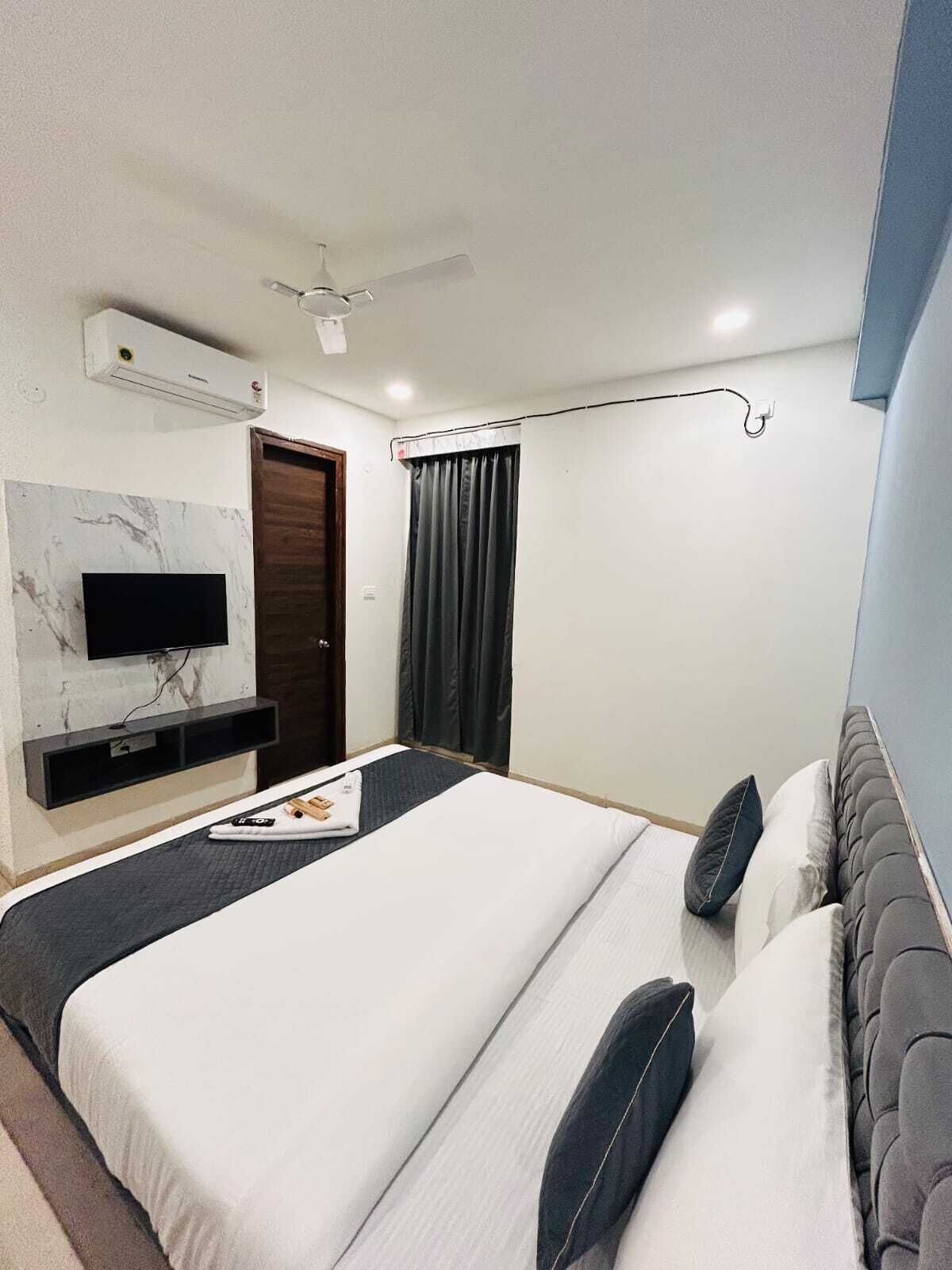 Una habitación de hotel moderna con televisión de pantalla plana montada en una pared de mármol, ventilador de techo, cama grande con ropa de cama oscura y clara, y ventana con cortinas oscuras.