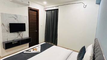 Una habitación de hotel moderna con televisión de pantalla plana montada en una pared de mármol, ventilador de techo, cama grande con ropa de cama oscura y clara, y ventana con cortinas oscuras.