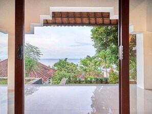 Vista desde una habitación con un ventanal grande, con vista a una piscina y un paisaje tropical con palmeras y cielo despejado.