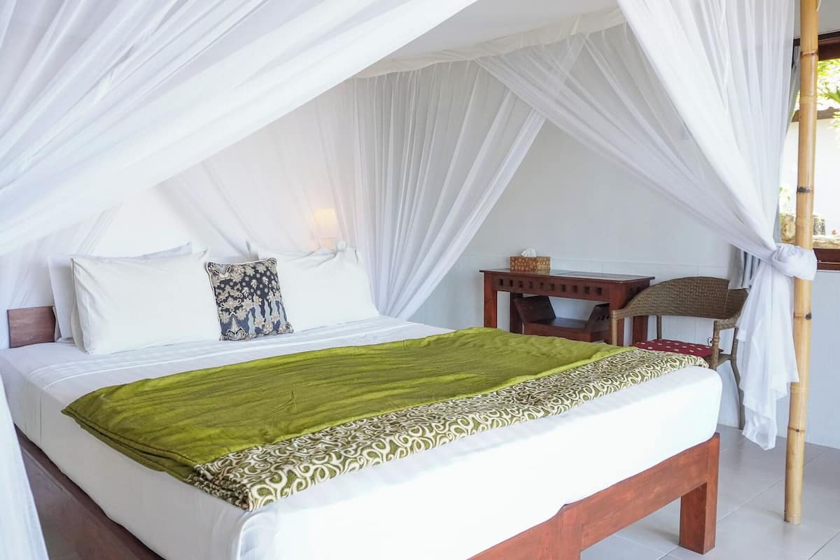 Cama com manta verde, almofadas brancas e um dossel. Tem uma mesinha de cabeceira de madeira com uma luminária e uma cadeira de vime.