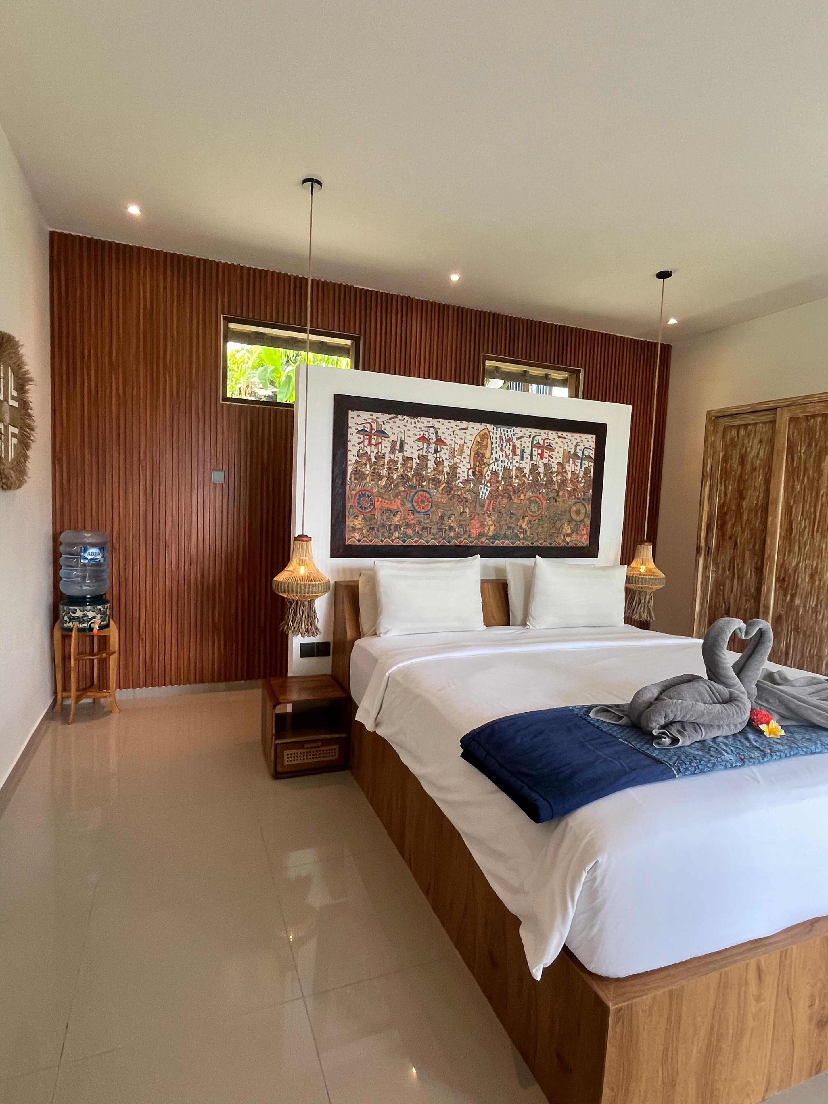 A hotel room with a large bed, wooden panel walls, a mounted artwork, a water dispenser, and a decorative swan pillow.