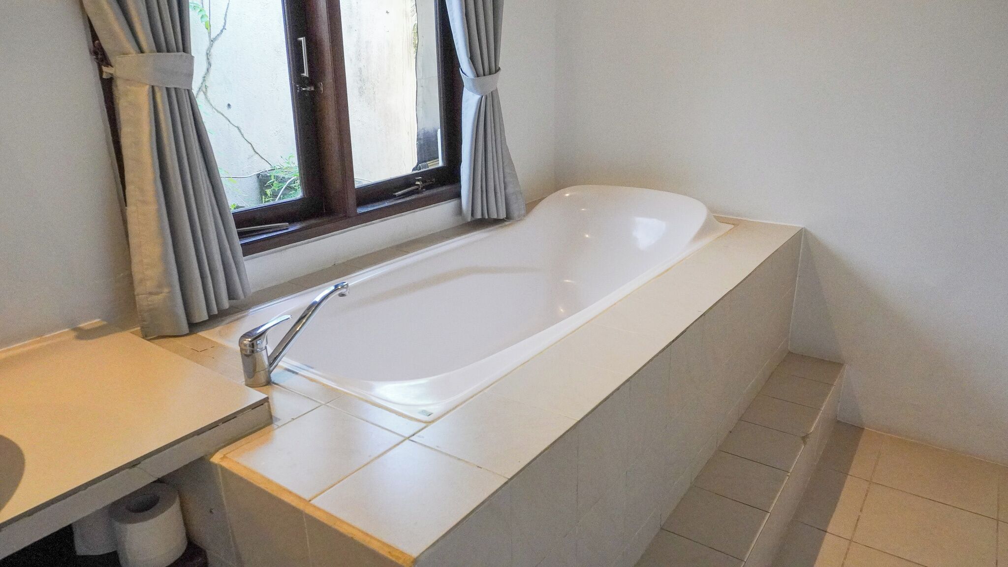 A bathroom with a white bathtub, a window with curtains, and a tiled floor.