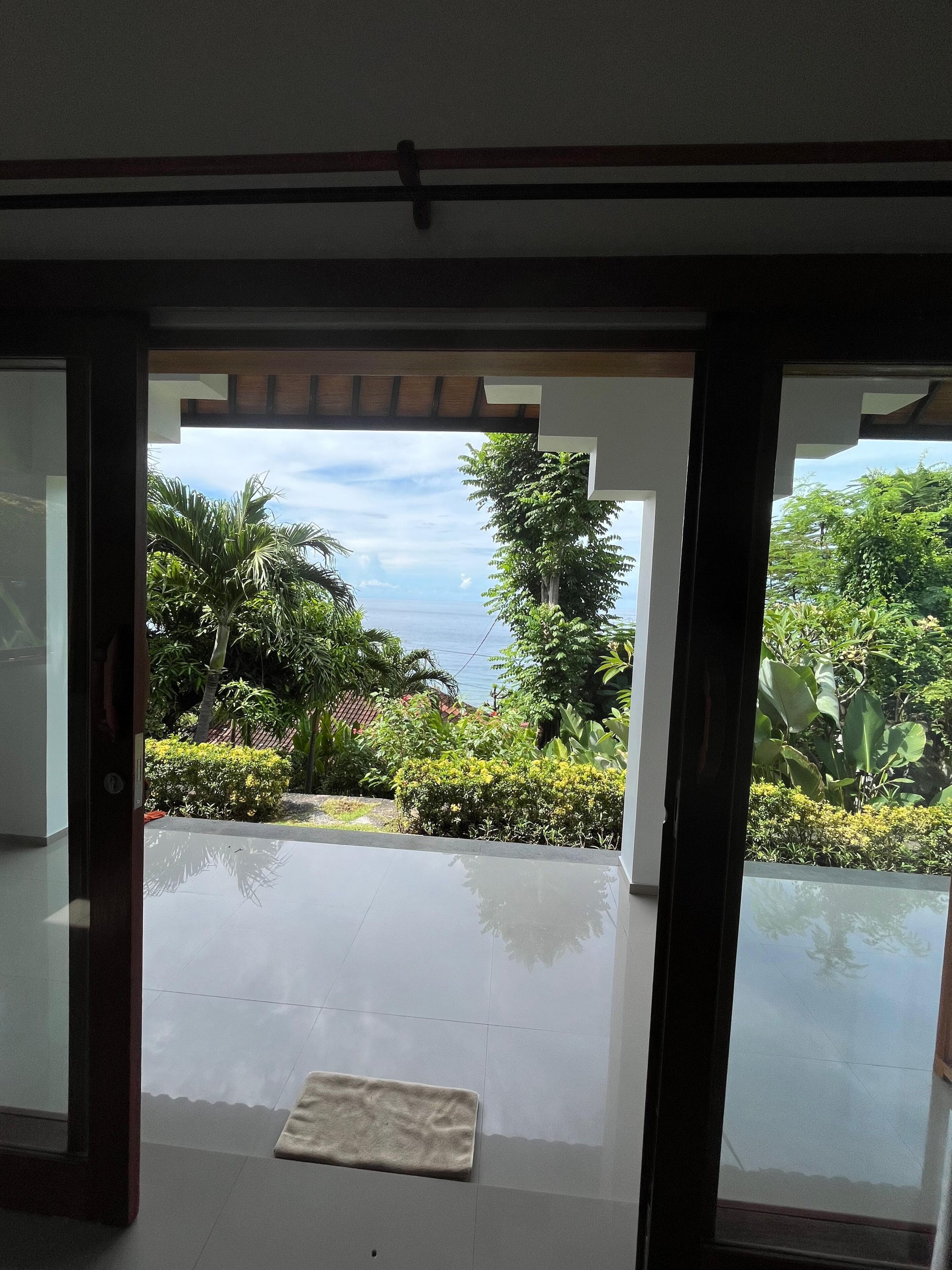 Une vue à travers une porte vitrée donnant sur un balcon, d’où l’on aperçoit un jardin tropical et la mer.