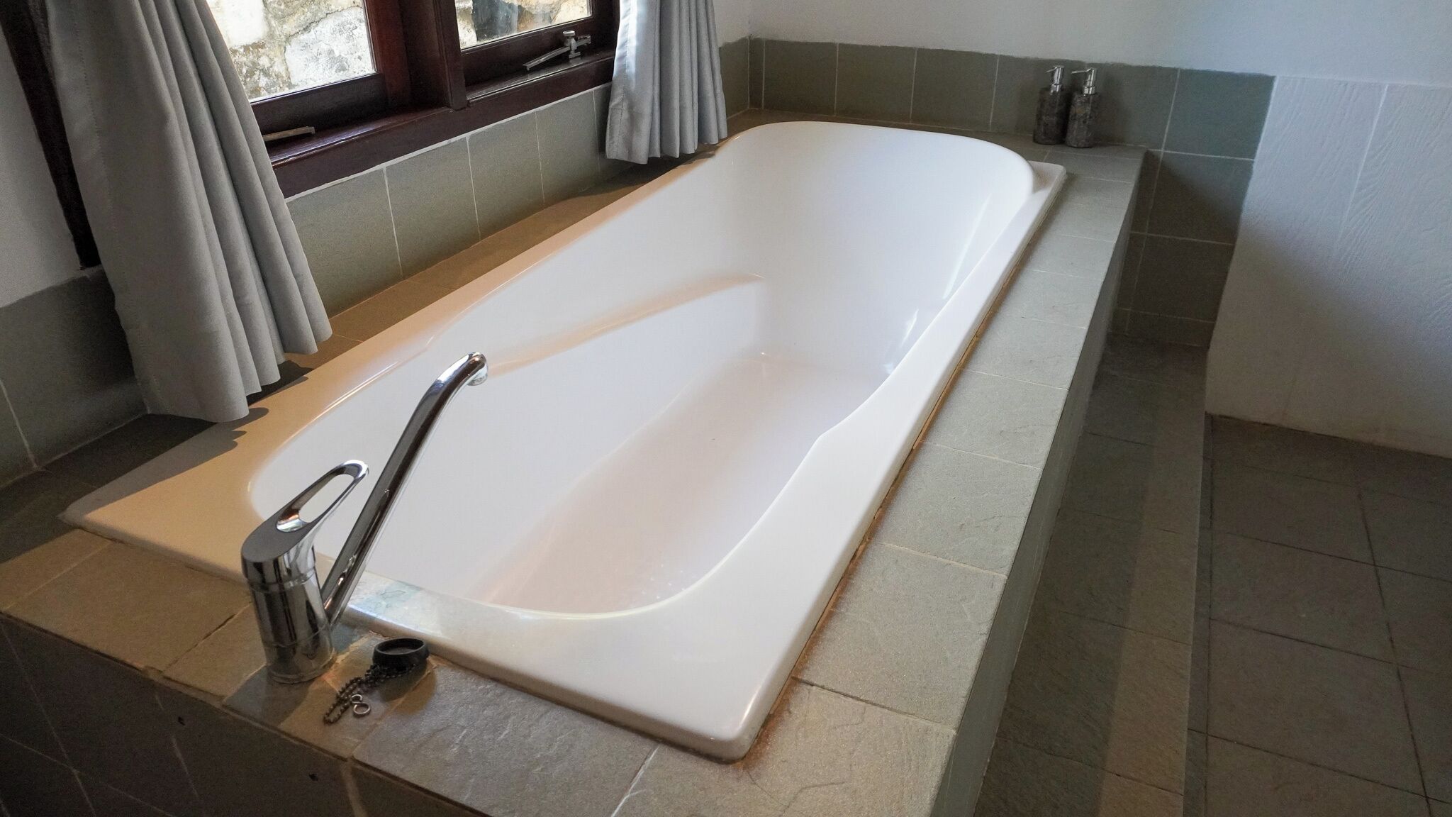 A modern bathroom with a freestanding bathtub, a window with curtains, and a tiled floor.