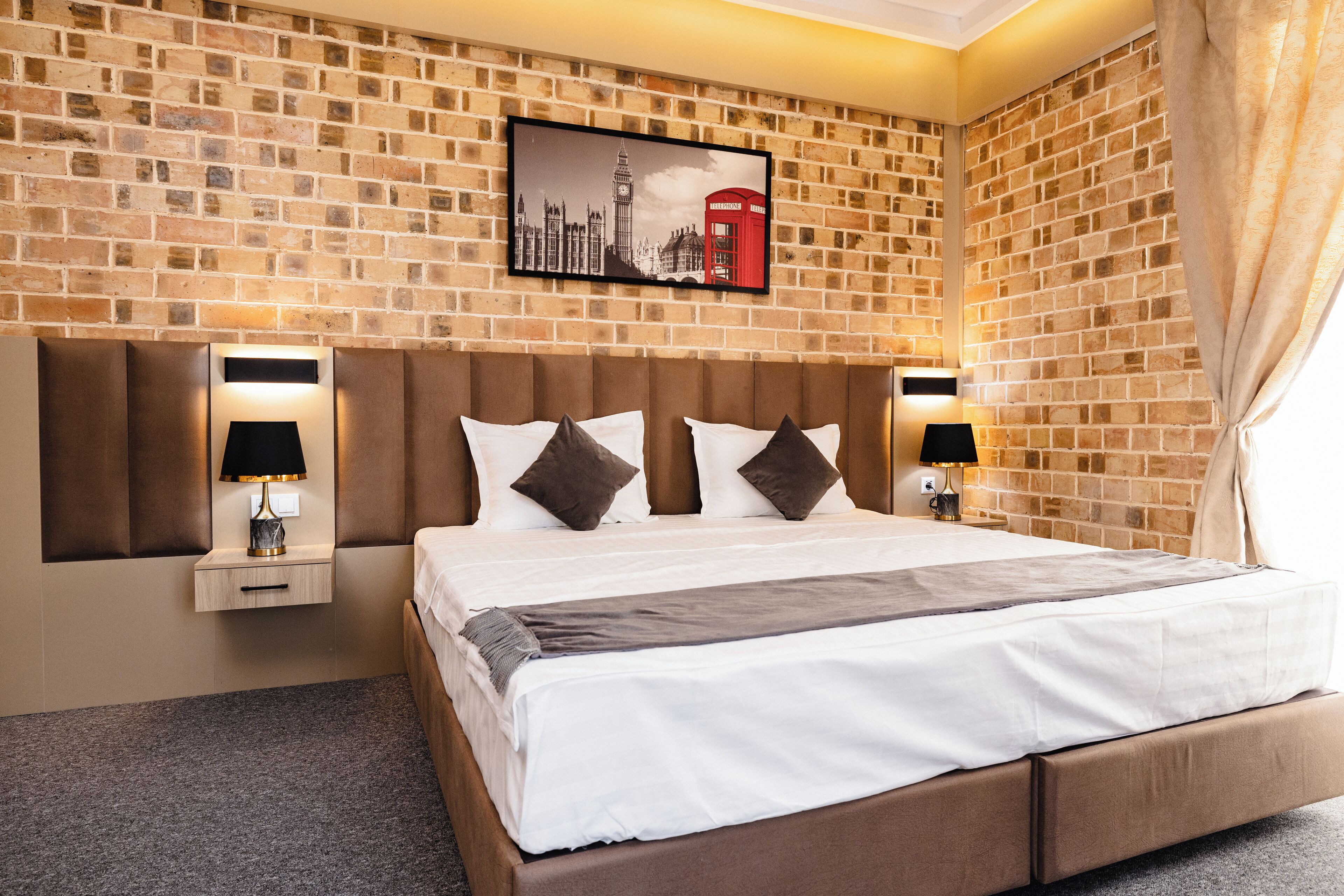 A hotel room with a brick wall, a large bed with a headboard, two bedside tables with lamps, and a framed picture of London landmarks.