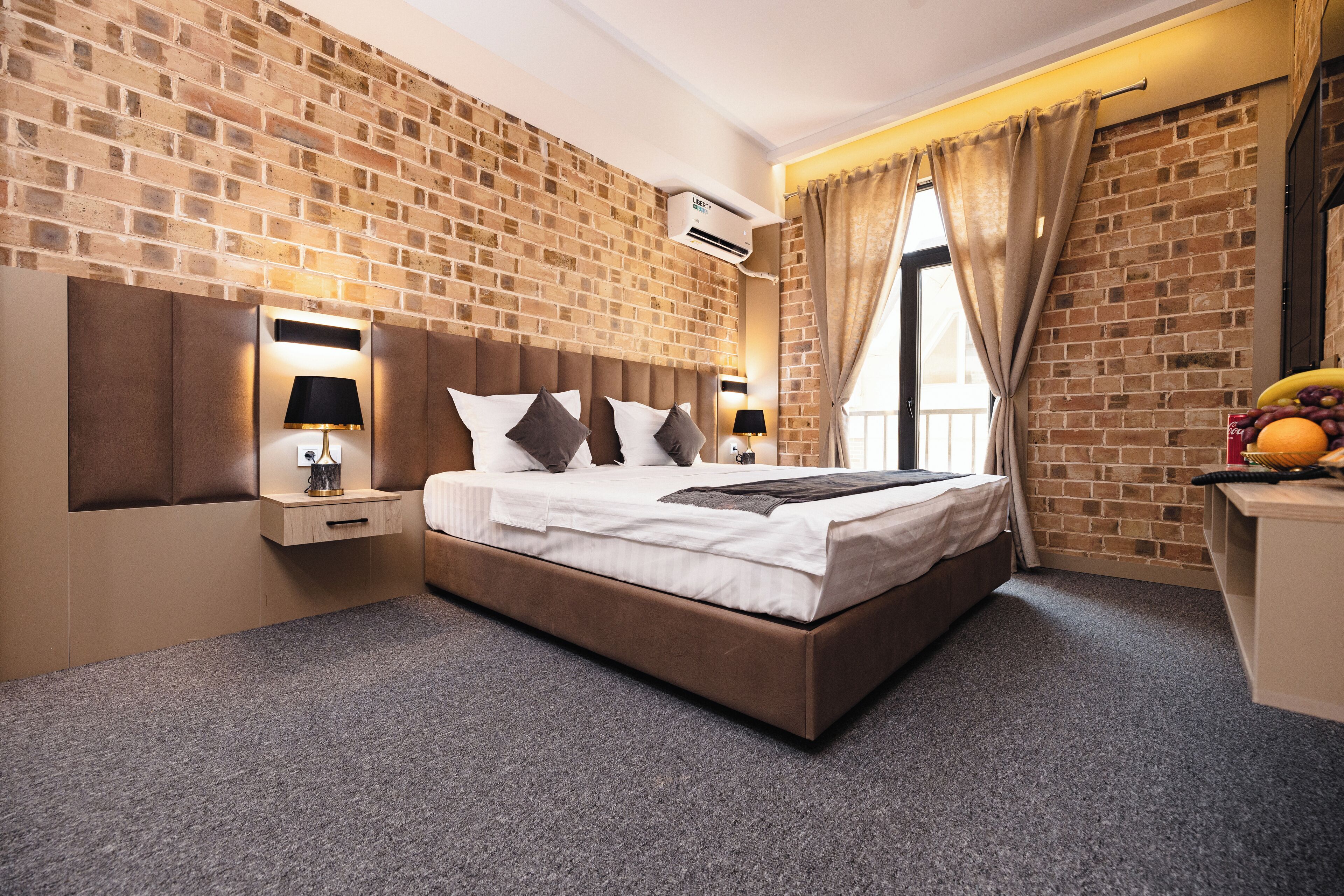 A modern hotel room with a brick wall, a large bed with white linens, two bedside tables with lamps, and a fruit bowl on a side table.