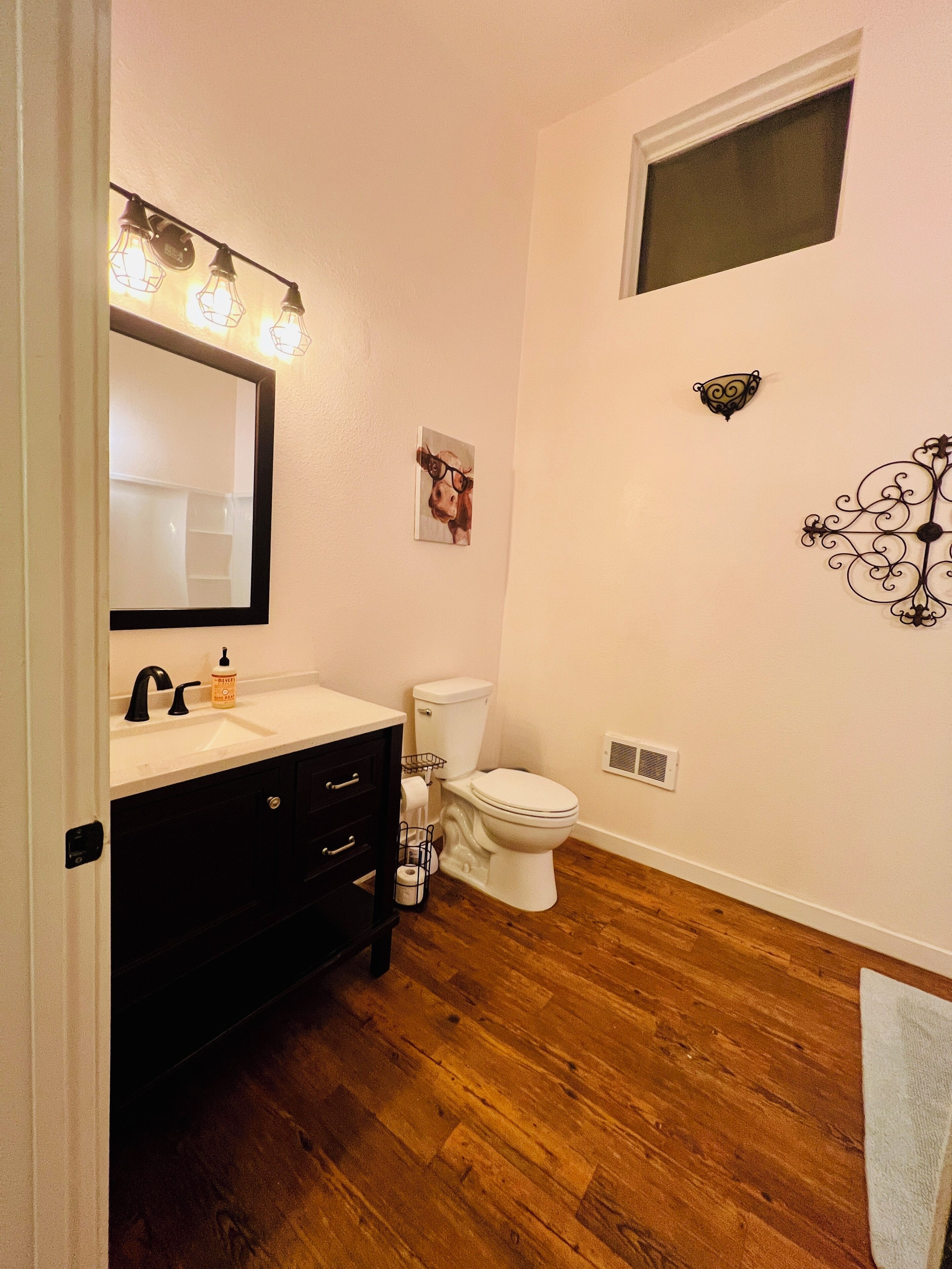Un baño con lavamanos blanco, tocador negro y inodoro blanco. Hay un espejo con luces sobre el lavamanos, una ventana pequeña sobre el inodoro y un adorno en la pared.