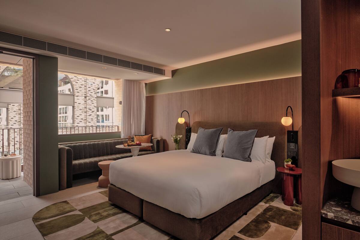 Ein modernes Hotelzimmer mit einem großen Bett, einer kleinen Sitzecke, einem Fenster mit Blick auf die Stadt und einem Badezimmer mit Spiegel und Waschbecken.