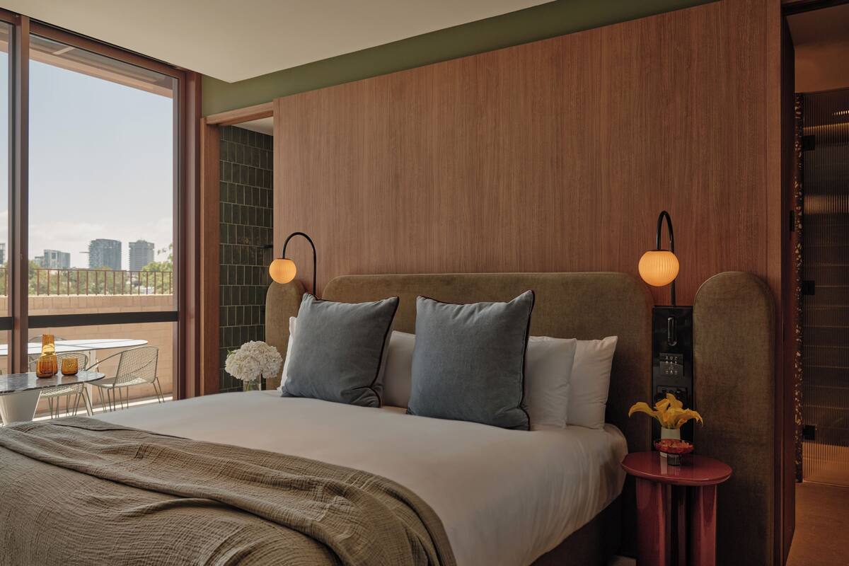 Ein modernes Hotelzimmer mit einem großen Bett, einem kleinen Tisch mit Blumen und Blick auf die Stadt durch große Fenster.