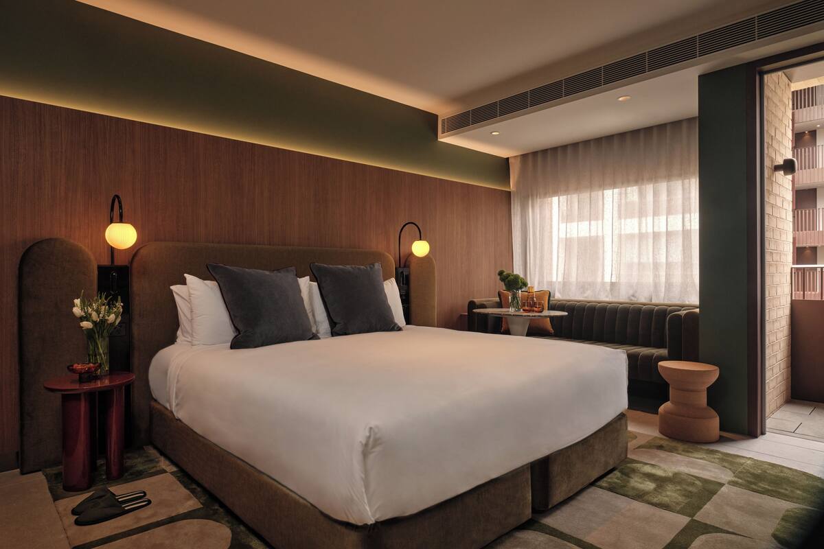 Ein modernes Hotelzimmer mit einem großen Bett, zwei Nachttischlampen, einem kleinen Tisch mit Blumen und Blick auf die Stadt durch große Fenster.