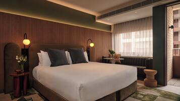 A modern hotel room with a large bed, two bedside lamps, a small table with a vase of flowers, and a view of the city through large windows.