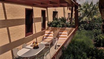 Une terrasse extérieure ensoleillée, avec un mur en briques, des coussins à rayures, une table garnie de fruits et des chaises en métal.
