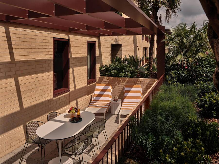 Une terrasse extérieure ensoleillée, avec un mur en briques, des coussins à rayures, une table garnie de fruits et des chaises en métal.