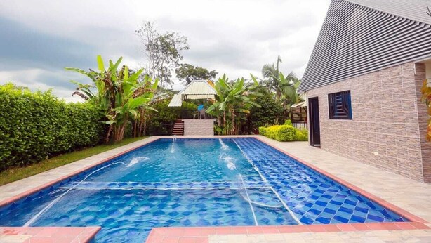 A swimming pool with a tiled edge, surrounded by greenery and a building with a sloped roof.
