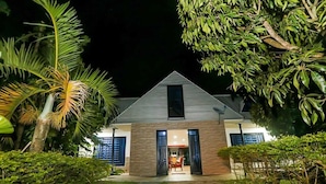 A modern two-story house with a triangular roof, surrounded by lush greenery and palm trees.
