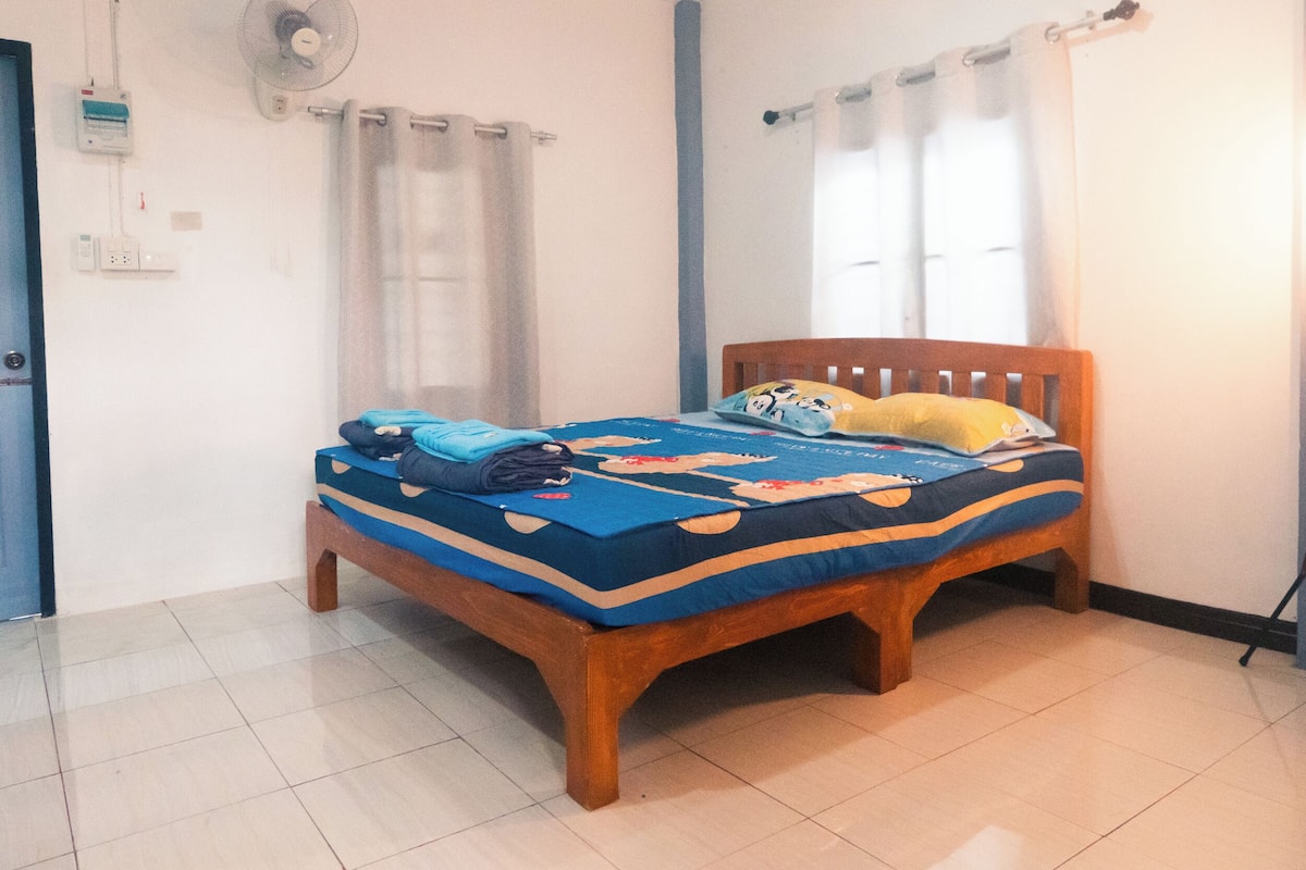 A bedroom with a wooden bed, a blue and orange bedspread, a fan, and a window with curtains.