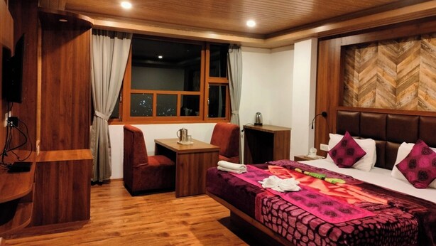 A hotel room with a wooden floor, a bed with a patterned bedspread, a desk with a chair, a window with curtains, and a wooden headboard. - Dragon Inn (Gangtok)