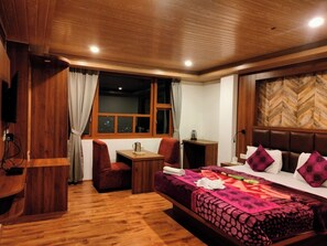 A hotel room with a wooden floor, a bed with a patterned bedspread, a desk with a chair, a window with curtains, and a wooden headboard. - Dragon Inn (Gangtok)