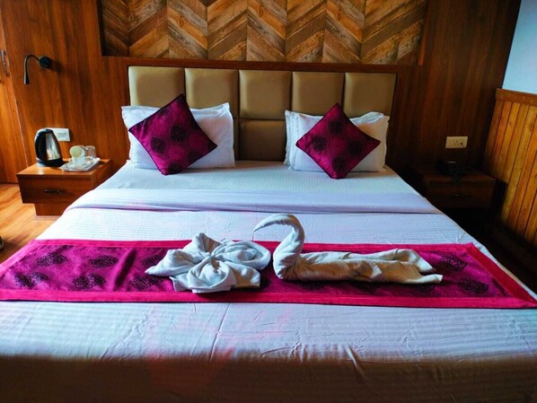 A hotel room with a large bed, two pillows, a nightstand with a kettle, and a red blanket with towels. - Dragon Inn (Gangtok)
