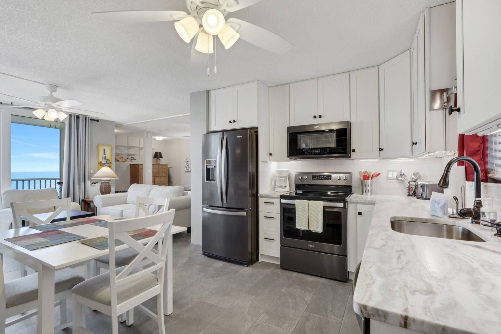 A modern kitchen with white cabinets, stainless steel appliances, and a large window overlooking the sea.