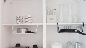 Een keuken met witte kastjes, twee roestvrijstalen pannen, een witte mok, een mok met dessin, een witte bak en een zwarte draadmand.