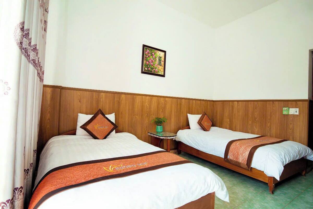 A hotel room with two beds, wooden headboards, a small table with a plant, and a framed picture on the wall.