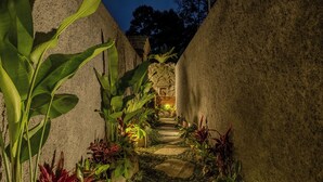 A stone pathway flanked by lush greenery and a stone wall, leading to a building with a warm light source. - Astala Ubud Villa (Ubud)
