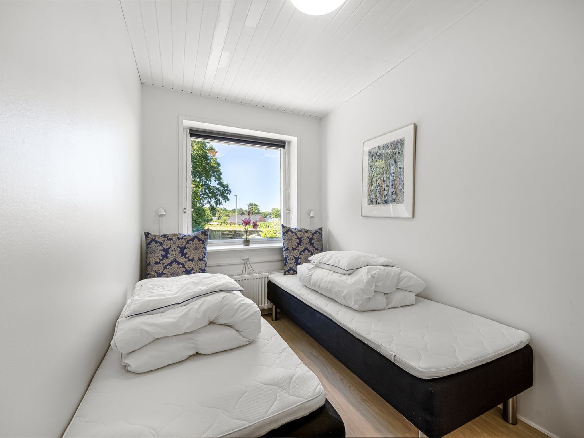 A small, bright room with two single beds, a window with a view of greenery, and a framed artwork on the wall.