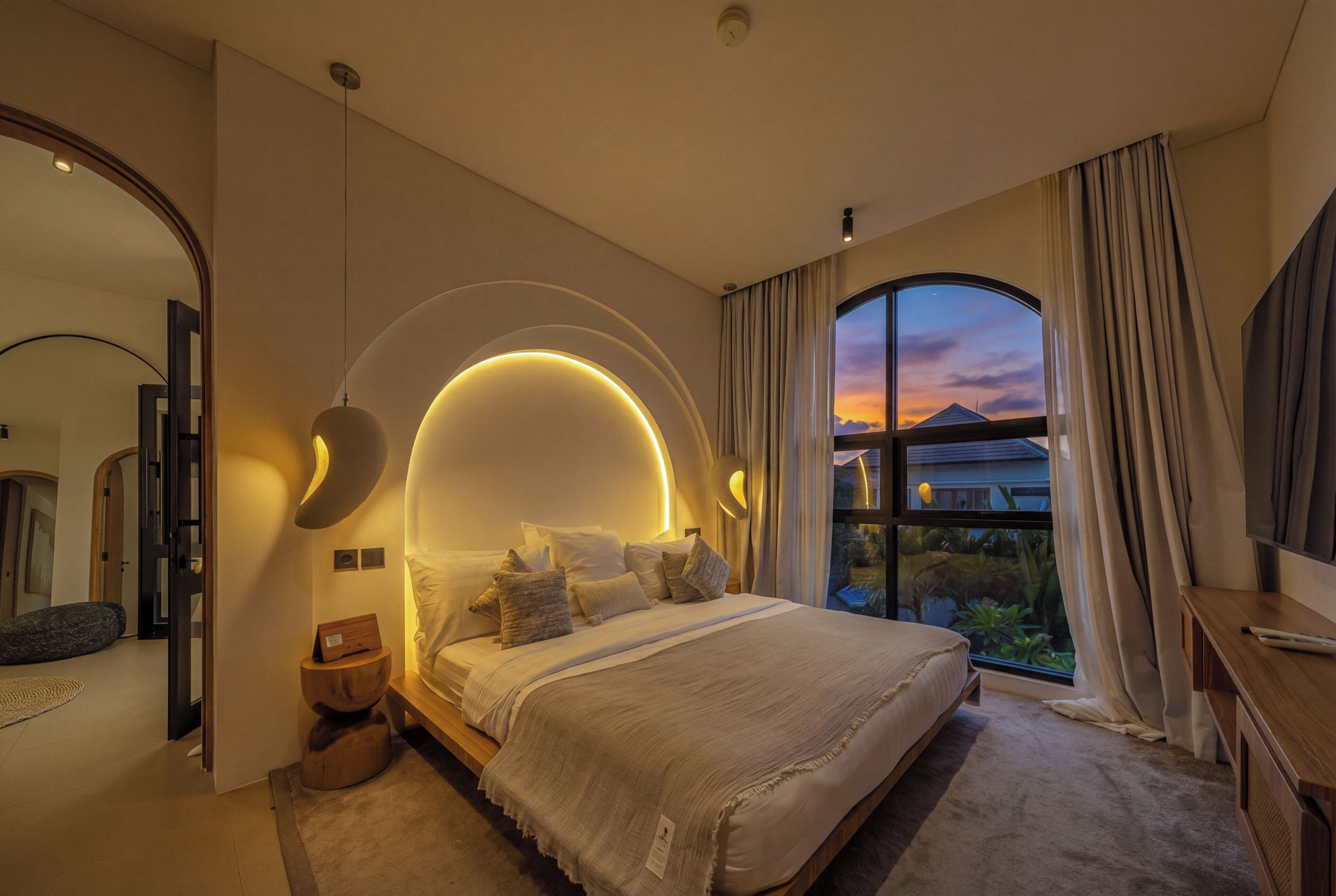 A modern bedroom with a large bed, a curved wall light, a large window with curtains, and a view of the outside.