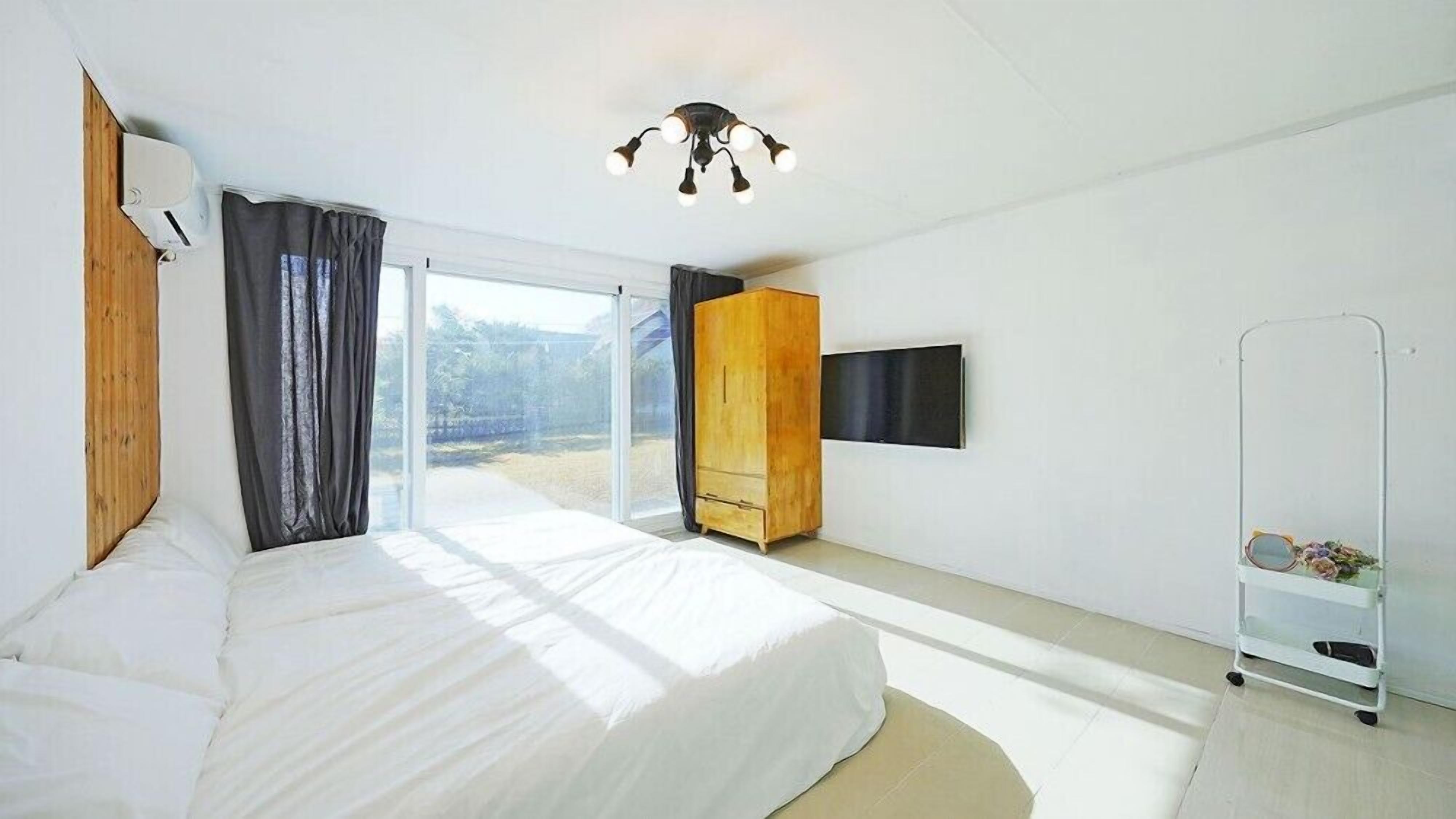 A modern bedroom with a large bed, a wardrobe, a TV mounted on the wall, and a sliding glass door leading to a balcony.