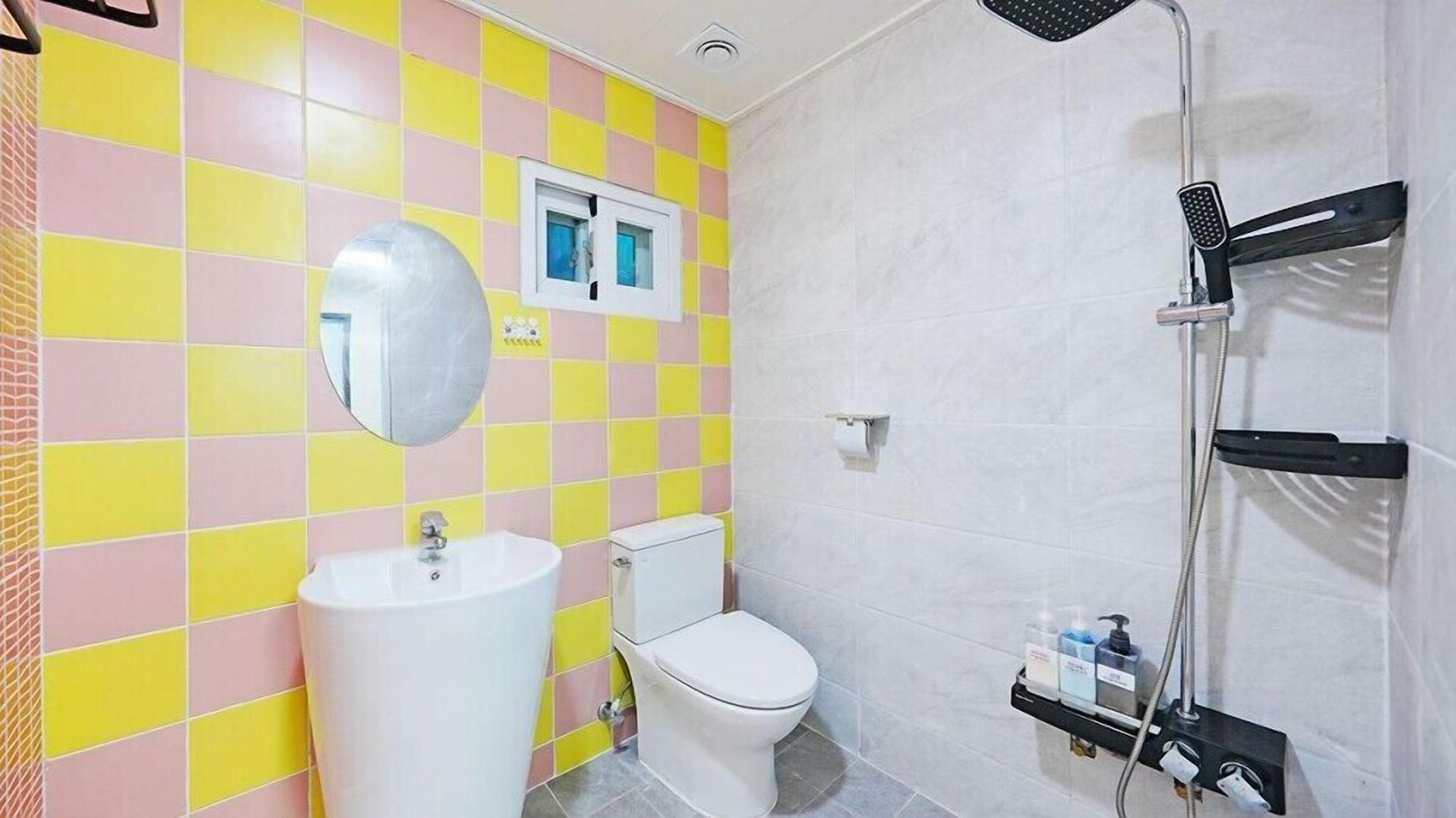 A bathroom with a white sink, toilet, and shower. The walls are tiled in a yellow and pink checkered pattern.