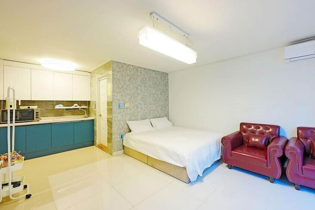 Une pièce de taille modeste comprenant un lit, un canapé en cuir rouge, une kitchenette et un luminaire blanc au plafond.