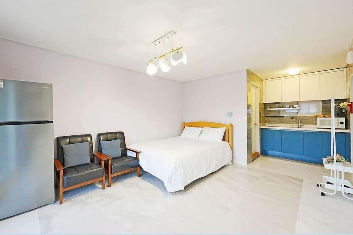 Une chambre d’hôtel de taille réduite, comprenant un lit, deux chaises, un réfrigérateur, un coin cuisine et un chariot de service.
