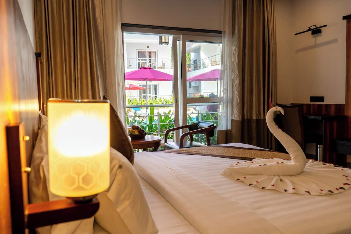 Habitación de hotel con cama, lámpara, almohada con forma de cisne, balcón con mesa y sillas, y vista a un edificio con balcones.