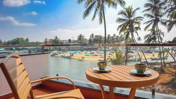 Un balcón con una silla y mesa de madera, con vista a una playa con barcos y palmeras.